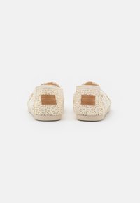 Zapatos tipo slip-on con un diseño de encaje en un color crema claro, que presentan un parche de cuero marrón claro con la etiqueta "TOMS" en el talón y una parte superior texturizada.