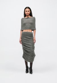 Femeie îmbrăcată cu un top scurt, off-shoulder, dintr-un material pufos de culoare gri închis și fustă midi asortată, colanți negri și tocuri negre cu bretele în jurul gleznei, stând împotriva unui fundal alb.