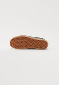 Semelle de chaussure avec motif gaufré en caoutchouc marron et bord en tissu vert, vue de dessous sur fond blanc.