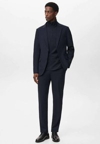 Completo blu con un pullover a collo alto, vestibilità sartoriale, tessuto liscio e discreta lucentezza. Scarpe eleganti nere completano l'outfit.