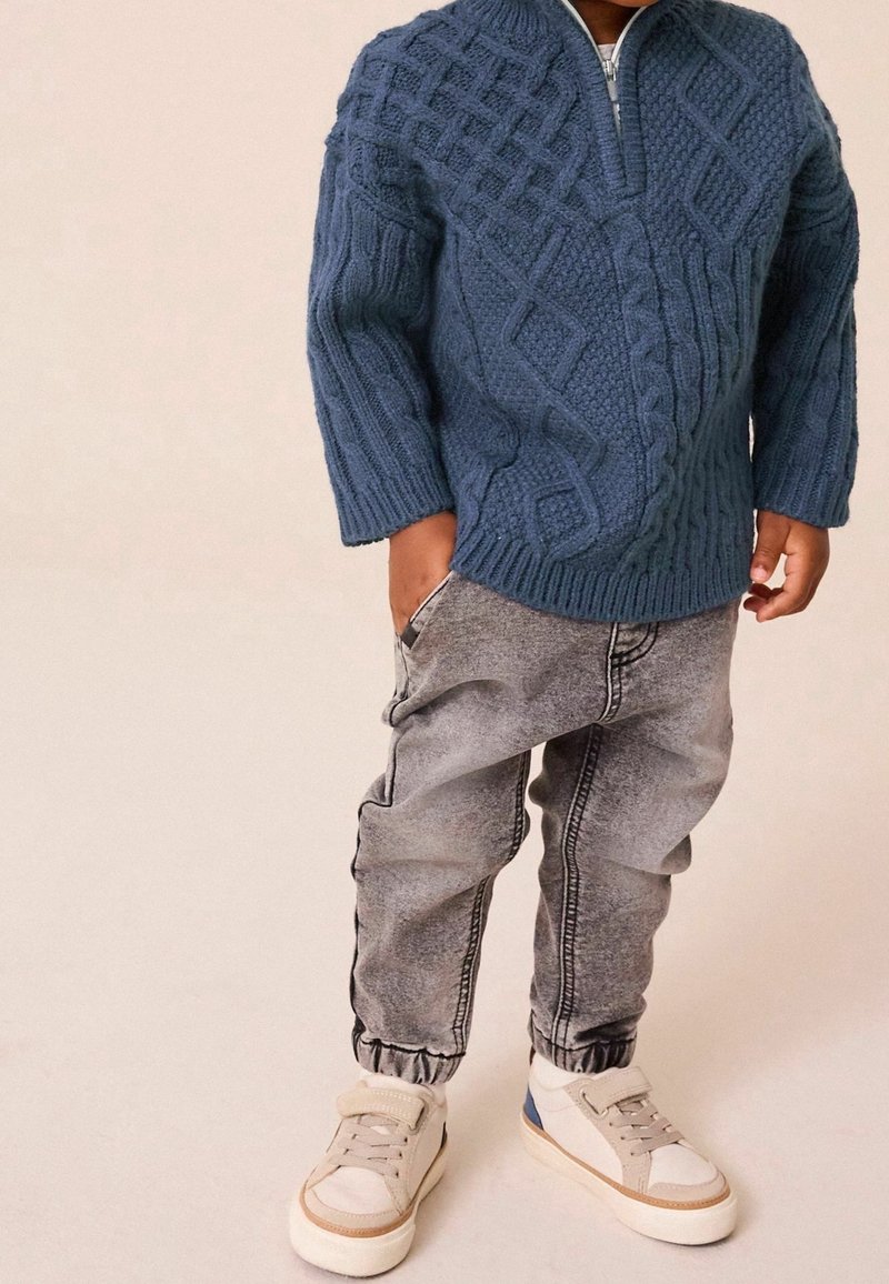 Enfant portant un pull en maille torsadée bleu, un jean gris délavé avec des poignets élastiques, et des baskets blanches avec des touches beige et bleu.