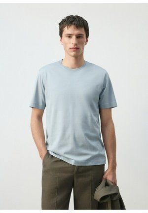 Jeune homme aux cheveux courts et foncés portant un t-shirt bleu clair et un pantalon marron, tenant une veste olive pliée dans la main droite, sur un fond uni.