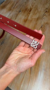 Red leather belt with silver "Hugo" logo buckle, featuring a smooth texture and rectangular shape, held in a hand against a wooden background.