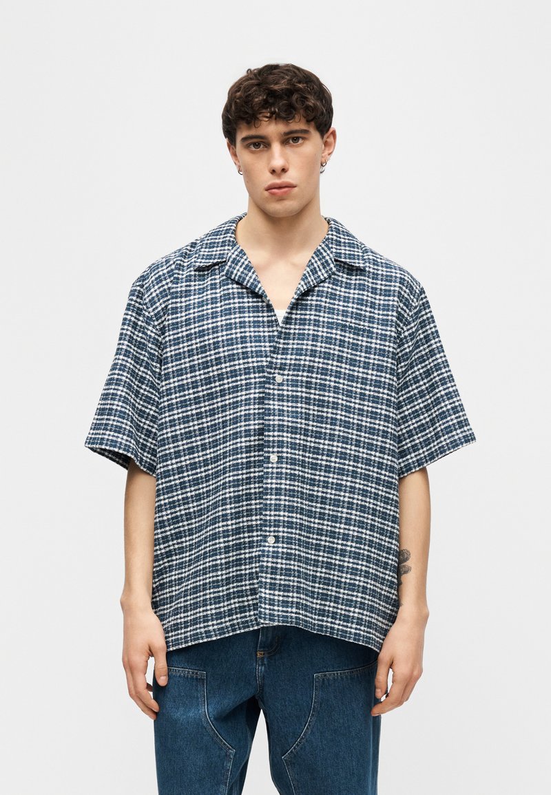 Joven con cabello corto y rizado, vestido con una camisa de manga corta de cuadros azul y blanca y jeans azules, posando frente a un fondo liso.