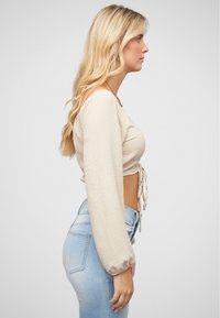 Crop top beige a maniche lunghe in tessuto testurizzato, con coulisse frontale e maniche a sbuffo, abbinato a jeans chiari a vita alta.