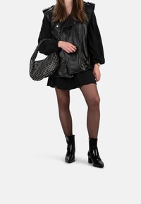 Gilet de motard en cuir noir sur une robe plissée noire, associé à des collants noirs transparents et des bottines brillantes. Comprend un sac à main noir clouté.
