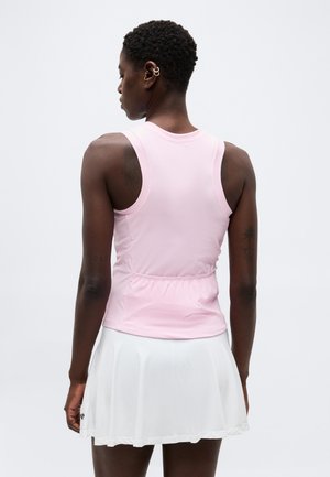 Γυναίκα μοντέλο με κοντά σγουρά μαλλιά που φορά ένα ροζ αμάνικο τοπ και λευκή πλισέ φούστα, φαινομένη από πίσω.