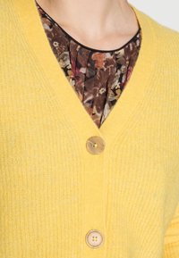 Cárdigan amarillo de punto con escote en V, tres botones y un sutil detalle texturizado. Superpuesto sobre una blusa de estampado floral con plisados.