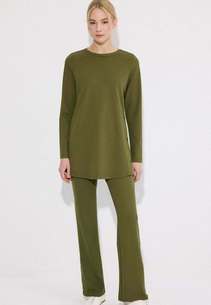 Kobieta stojąca, ubrana w oliwkowo-zieloną tunikę z długim rękawem oraz dopasowane spodnie o prostych nogawkach, w białych tenisówkach na białym tle.