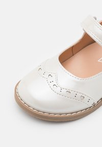 White patent leather Mary Jane shoe with heart cutouts along the scalloped edge, brown rubber sole, and adjustable strap.