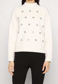 Žena v krémovém svetru s opakovaným černým vzorem loga "KL" a černých žebrovaných kalhotách, stojící na jednobarevném pozadí.