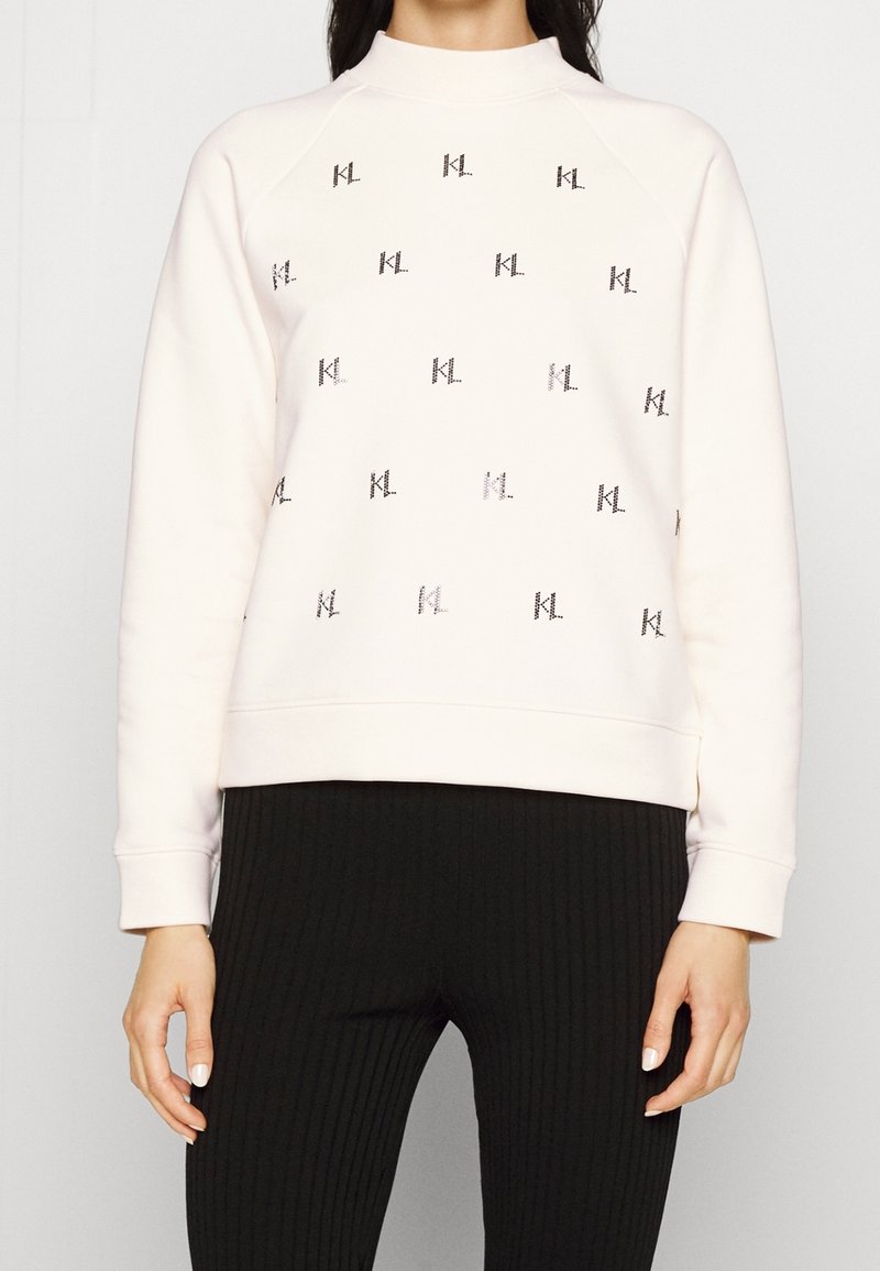 Žena v krémovém svetru s opakovaným černým vzorem loga "KL" a černých žebrovaných kalhotách, stojící na jednobarevném pozadí.
