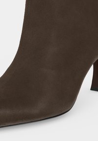 Bout de bottine en daim marron avec une forme carrée arrondie et un petit talon carré visible à l’arrière sur fond blanc.