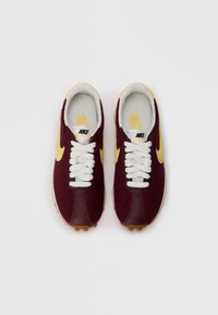 Bordo Nike sportbačiai su geltonais akcentais, baltais raiščiais ir tekstūruota viršumi. Yra guminis padas su šviesiai rudais protektoriais, užtikrinančiais sukibimą.