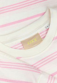 Υφάσμα με άσπρες και ροζ ρίγες, με επικαλυπτόμενο λαιμόκοψη και μπεζ ετικέτα ρούχου που γράφει «JJXX» με κίτρινα γράμματα.