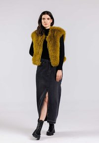 Faux-Fur-Weste in Senfgelb, kombiniert mit einem schwarzen langärmligen Oberteil und einem dunkelblauen Jeansrock mit einem Schlitz vorne. Schwarze Schnürstiefel runden den Look ab.