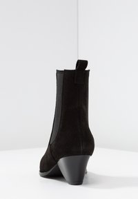 Botas de ante negras de tobillo con un tacón afilado, paneles laterales elásticos y una lengüeta en la parte posterior para facilitar su puesta. Textura suave.