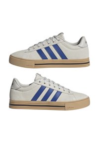 Sneaker Adidas con tomaia in suede grigio chiaro, tre strisce blu e suola in gomma color caramello con linee di accento orizzontali.