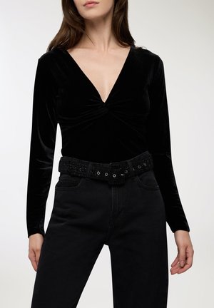 Body in velluto nero con maniche lunghe, scollo a V e dettaglio frontale attorcigliato, abbinato a jeans neri a vita alta e a una cintura nera strutturata.
