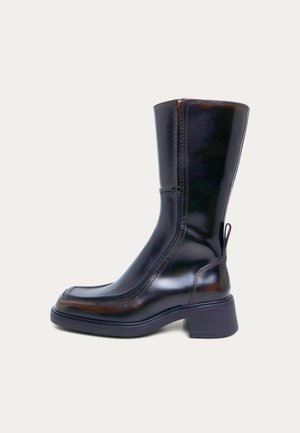 Botte noire brillante en cuir, montant jusqu’au genou, avec des empiècements cousus, un bout arrondi et un talon bas et épais, sur fond uni.
