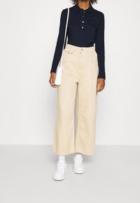 Navy long-sleeve polo shirt with button placket, paired with light beige wide-leg trousers and white sneakers. Minimalistic white crossbody bag.