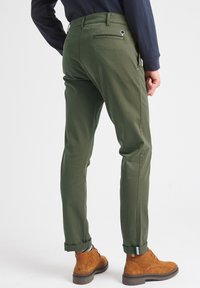 Chinos de algodón verde con un ajuste slim, con un bolsillo trasero con cremallera y puños enrollados, combinados con botines de ante marrón.