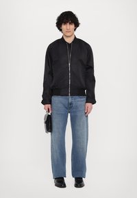 Zwarte bomberjack met een ritssluiting aan de voorkant, gecombineerd met lichtblauwe rechte jeans. Het model houdt een zwarte tas vast en draagt zwarte schoenen.