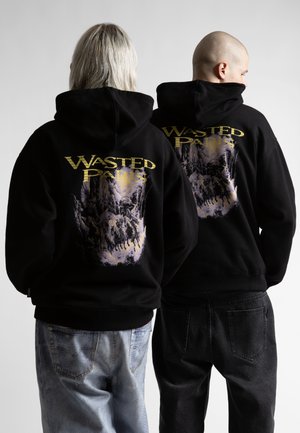 Dvaja ľudia stoja chrbtom ku kamere, oblečení v čiernych mikinách s kapucňou s nápisom „WASTED PARIS“ a grafikou koní a jazdcov v hmlistom lese.
