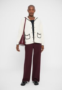 Cárdigan blanco con canalé y ribete negro, combinado con pantalones de pierna ancha en burdeos oscuro y un bolso burdeos. Zapatos negros con costuras decorativas.