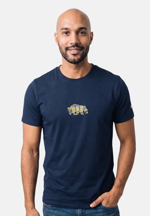 Homme avec barbe et tête rasée portant un t-shirt bleu marine avec un petit motif abstrait jaune sur la poitrine, debout devant un fond blanc.