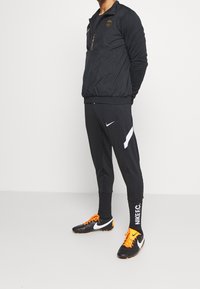Sort træningsjakke med guld logo, halv-lynlås og elastisk kant. Paireret med sorte bukser med hvid stribe og orange sportssko.