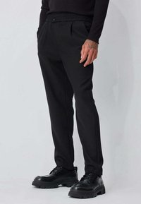 Pantalon noir ample avec taille élastique et fermeture par bouton, doté de poches latérales et d'un tissu lisse. Associé à des chaussures noires épaisses.