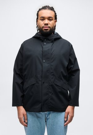 Mand med fletninger i håret og skæg, iført en sort hættetrøje og lyseblå jeans, stående mod en ensartet hvid baggrund.