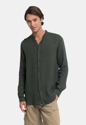 Giovane uomo con capelli scuri di media lunghezza, indossa una camicia verde scuro abbottonata e pantaloni cargo beige, in piedi davanti a uno sfondo semplice.