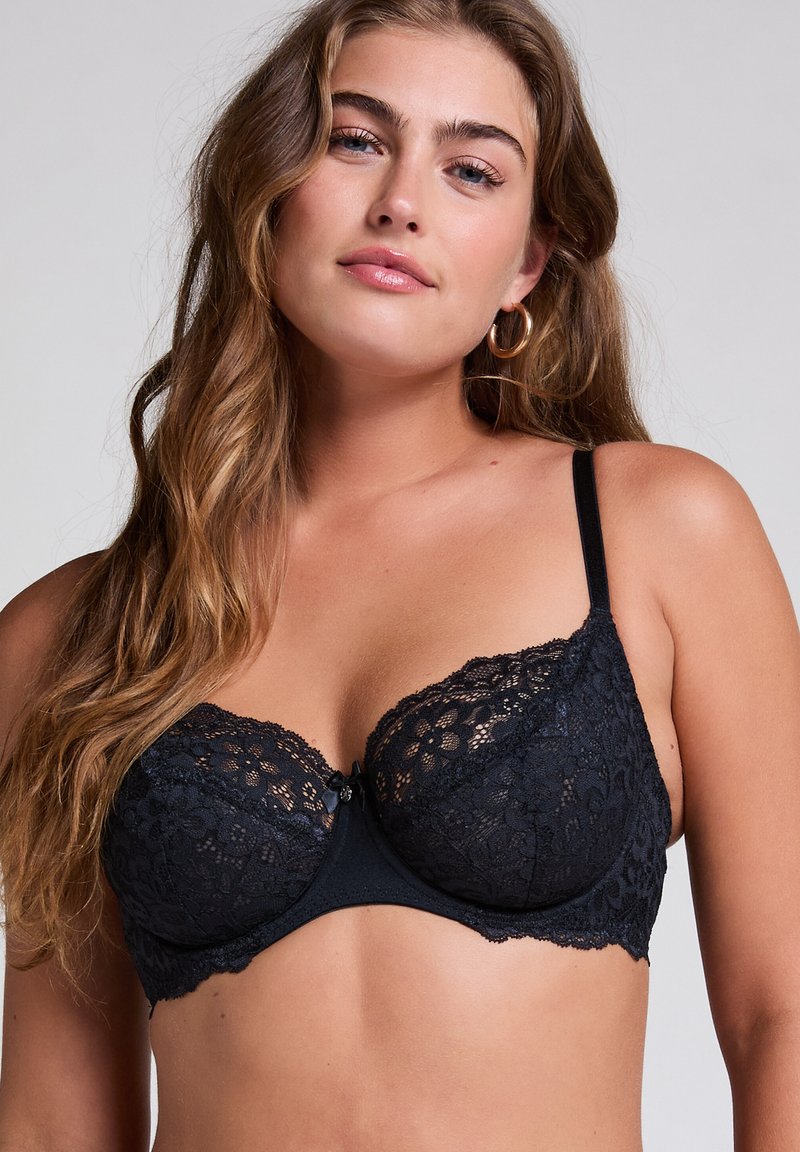 Nő hosszú hullámos hajjal, arany karika fülbevalóval és fekete csipkés melltartóval, világos, egyszínű háttér előtt áll.