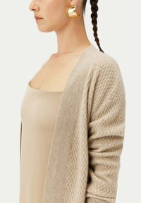 Beige stickad cardigan med ett texturerat mönster, öppen framtill och långa ärmar, tillsammans med en neutral tanktop.