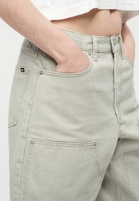 Άτομο που φοράει ανοιχτό γκρι τζιν με τα χέρια στις μπροστινές τσέπες, εμφανή μέση και λευκό κοντό τοπ.