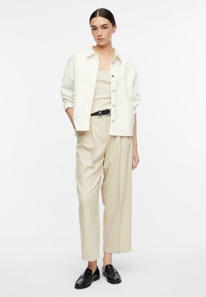 Donna che indossa una giacca bianca con bottoni, canottiera beige e pantaloni a gamba larga, cintura nera e mocassini neri, in piedi con le mani parzialmente nelle tasche.