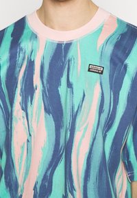 Tričko Adidas s vlnkovým vzorem v aqua, námořnické a růžové barvě. Klasický kulatý výstřih s prošitým lemem a malou značkovou nášivkou.