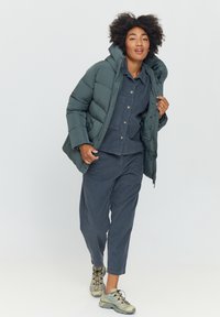 Dark green puffer jacket with a high collar, paired with a blue corduroy shirt and matching pants. Beige sporty shoes with black laces.