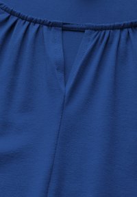 Niebieska bluzka z marszczonym dekoltem, otwarciem w kształcie litery V oraz gładkim, lekkim materiałem o teksturowanym wykończeniu.