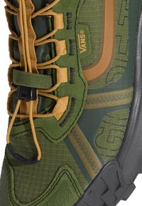 Gros plan sur une basket Vans verte et beige montrant un tissu texturé, des parties rembourrées, des lacets beiges avec un bouton noir et des détails de semelle en caoutchouc noire.