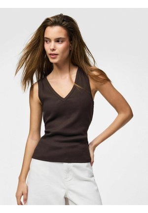 Žena s dlhými vlasmi, ktorá má na sebe tmavohnedý tielkový top s výstrihom do V a biele nohavice, stojaca s jednou rukou na boku.