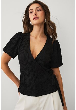 Kobieta ubrana w czarny, teksturowany, kopertowy top z bufkami na rękawach oraz białe spodnie, pozująca na jasnym, jednolitym tle.