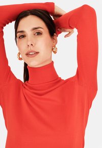 Červený rolák vyrobený z měkkého materiálu, s dlouhými rukávy a žebrovanou texturou. Velké zlaté kruhové náušnice doplňují design.