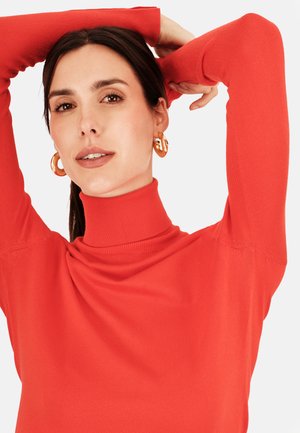 Červený rolák vyrobený z měkkého materiálu, s dlouhými rukávy a žebrovanou texturou. Velké zlaté kruhové náušnice doplňují design.