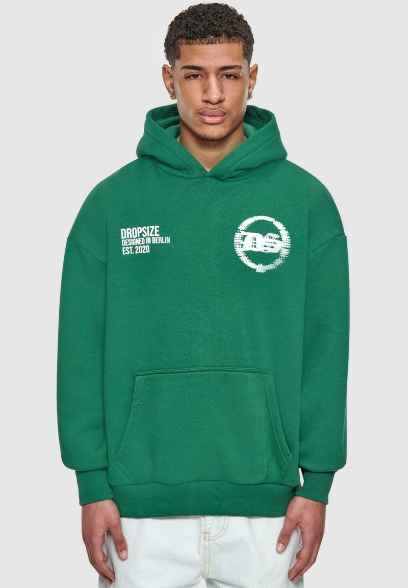 Hoodie vert avec une poche avant, orné de texte imprimé blanc et d'un logo circulaire sur le côté gauche. Tissu doux, coupe décontractée.