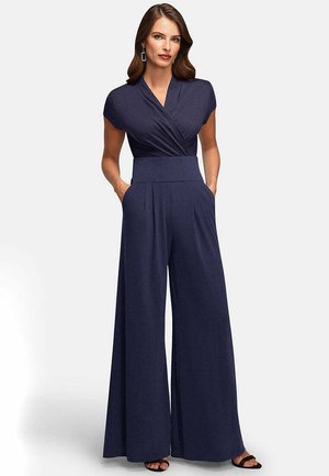 Žena oblečená v námornícky modrom širokonohom overal s krátkymi rukávmi, hlbokým výstrihom do V, vysokým pásom a rukami v vreckách, stojaca pred jednoduchým pozadím.
