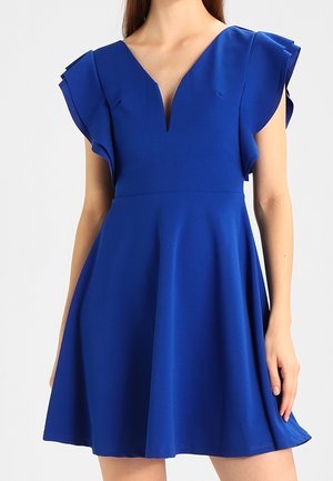 Vrouw draagt een felblauwe jurk tot op de knie met fladdermouwen en een diep V-hals tegen een effen achtergrond.
