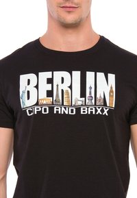 Schwarzes T-Shirt mit dem Wort "BERLIN" in großen weißen Buchstaben und verschiedenen Illustrationen von Sehenswürdigkeiten darunter. Die Markenbezeichnung "CIPO AND BAXX" ist ebenfalls enthalten.